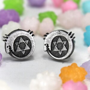 May include: A pair of silver earrings featuring a black and white symbol with a star and a crescent moon. The symbol is surrounded by a circle with a jagged edge.