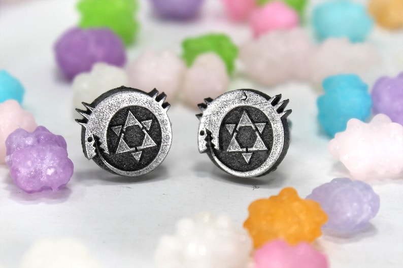 May include: A pair of silver earrings featuring a black and white symbol with a star and a crescent moon. The symbol is surrounded by a circle with a jagged edge.