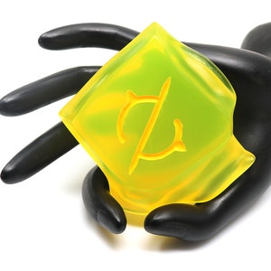May include: A translucent, yellow-green, diamond-shaped object with a stylized "C" design in the center. The object is held by a black hand against a white background. The object appears to be made of a clear, possibly plastic material.