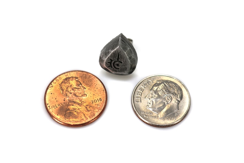 May include: A silver metal die with a symbol on each side. The die is about the size of a US penny.