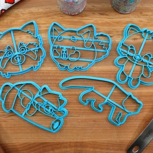 May include: Five blue plastic cookie cutters shaped like red pandas. The cutters are on a wooden surface.
