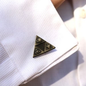 May include: Gold-toned cufflinks featuring a triangular design with a stylized image of a sword, a heart, and a star.