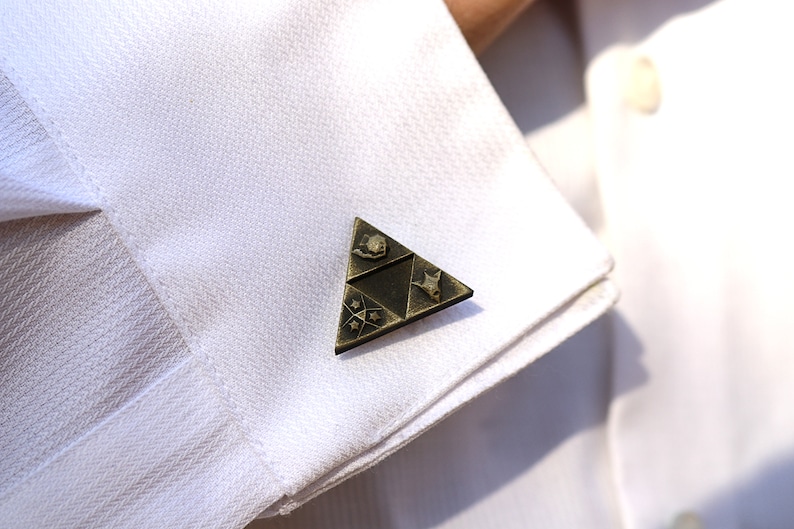 May include: Gold-toned cufflinks featuring a triangular design with a stylized image of a sword, a heart, and a star.
