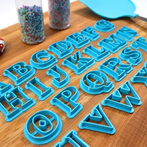 May include: A set of blue plastic alphabet cookie cutters on a wooden cutting board. The cutters are arranged in alphabetical order.