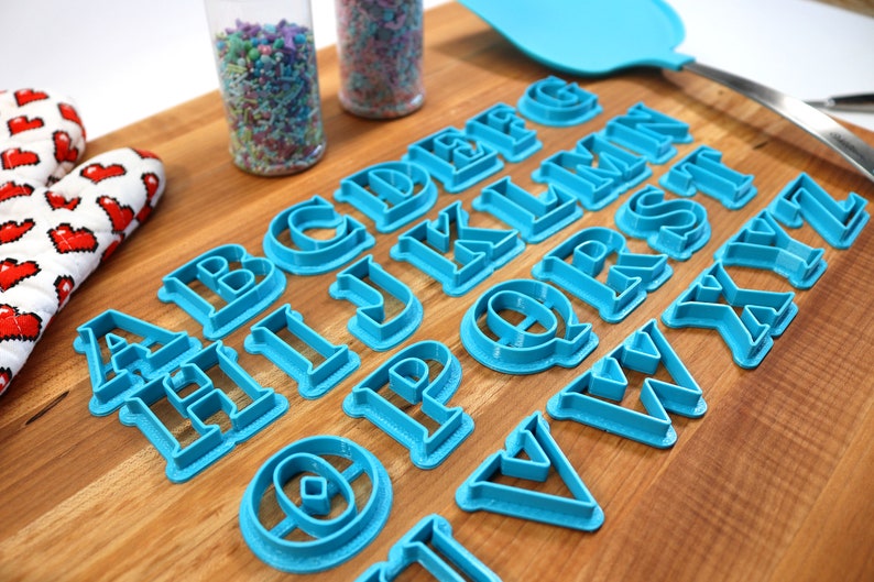 May include: A set of blue plastic alphabet cookie cutters on a wooden cutting board. The cutters are arranged in alphabetical order.