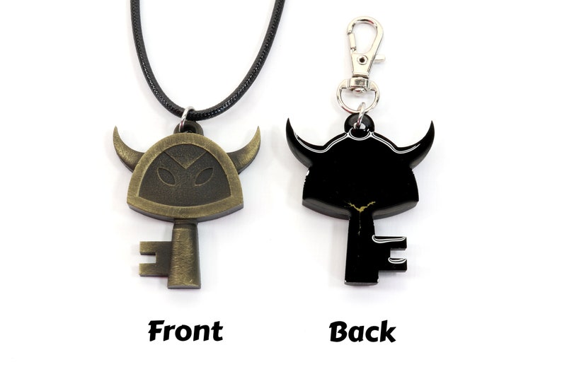 May include: A gold-colored key pendant with a horned helmet design. The key has a black silhouette on the back. The pendant is on a black cord and a silver key chain.