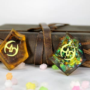 May include: Two resin bookmarks with a gold symbol and glitter embedded in the resin. The bookmark on the left is amber colored and the bookmark on the right is green and gold.