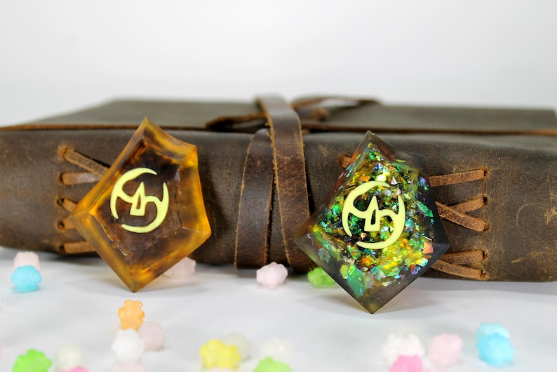 May include: Two resin bookmarks with a gold symbol and glitter embedded in the resin. The bookmark on the left is amber colored and the bookmark on the right is green and gold.