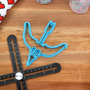 May include: A blue plastic cookie cutter shaped like a crescent moon with an arrow through it. The cutter is on a wooden surface with a black ruler.