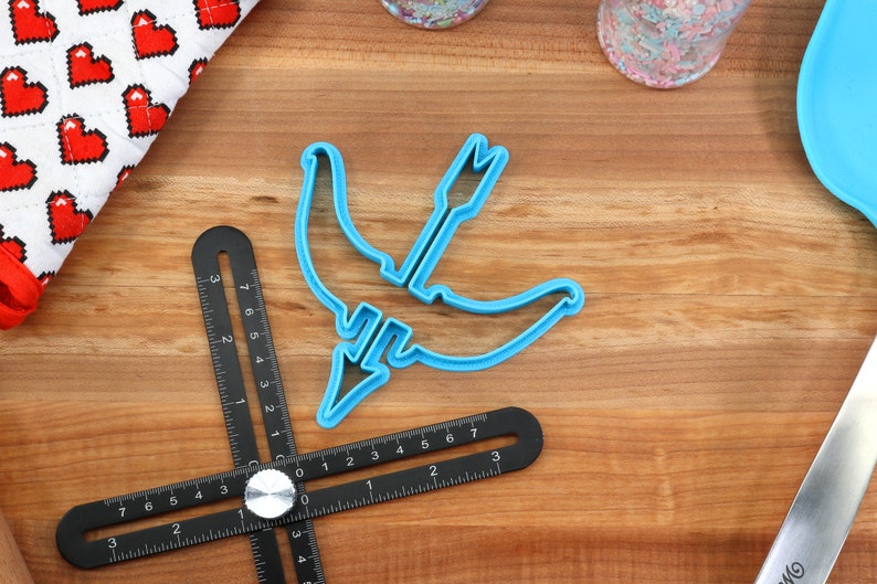 May include: A blue plastic cookie cutter shaped like a crescent moon with an arrow through it. The cutter is on a wooden surface with a black ruler.