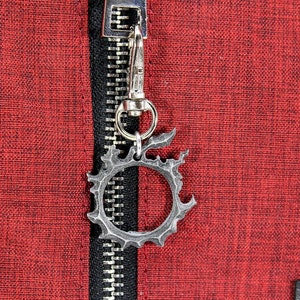 Puede incluir: Un llavero de metal plateado con un dise&ntilde;o circular con borde dentado. El llavero est&aacute; unido a una cremallera en una bolsa de tela roja.