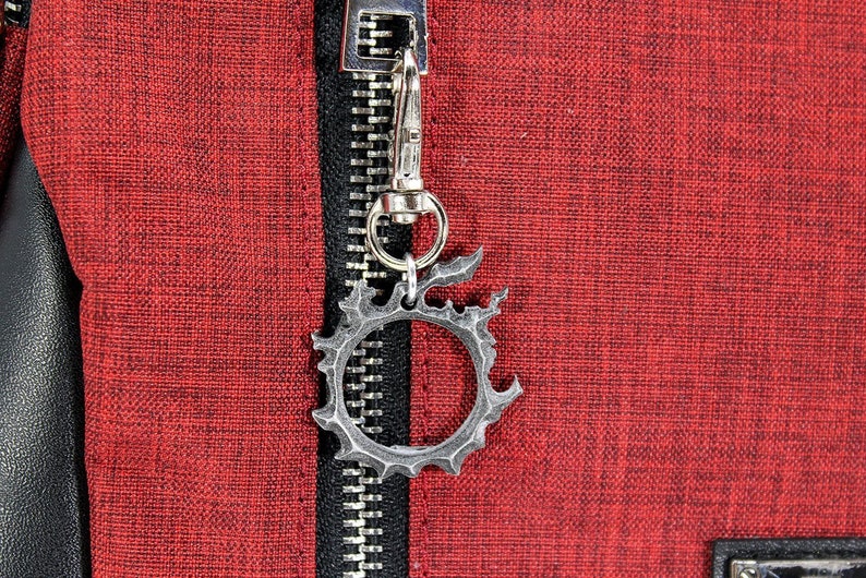 Puede incluir: Un llavero de metal plateado con un dise&ntilde;o circular con borde dentado. El llavero est&aacute; unido a una cremallera en una bolsa de tela roja.