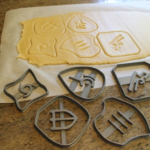 May include: A set of six gray plastic cookie cutters with different designs, including a shield, a football, and a swirl. The cutters are on a white surface with a sheet of rolled out cookie dough in the background.