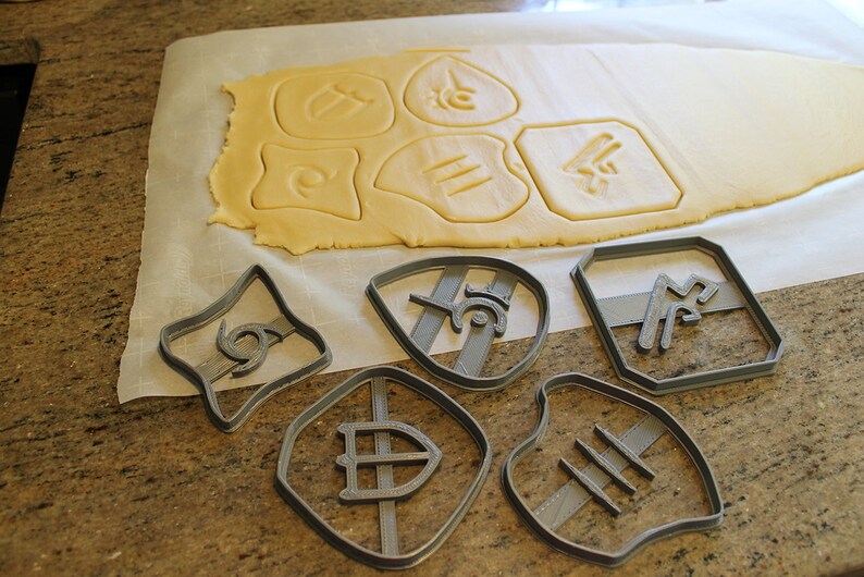 May include: A set of six gray plastic cookie cutters with different designs, including a shield, a football, and a swirl. The cutters are on a white surface with a sheet of rolled out cookie dough in the background.