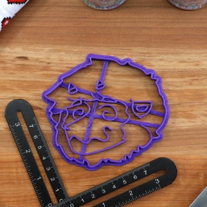 May include: Purple plastic cookie cutter shaped like a cartoon face with a wide smile and large eyes.