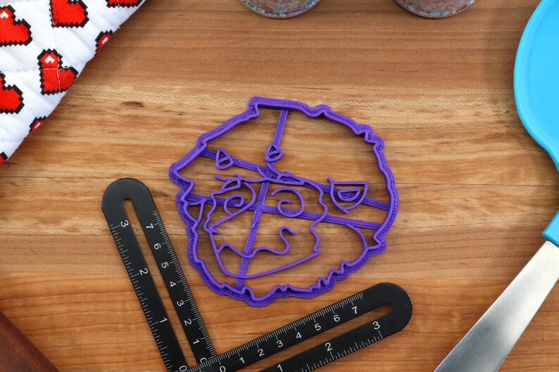 May include: Purple plastic cookie cutter shaped like a cartoon face with a wide smile and large eyes.