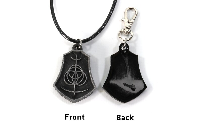 May include: A black leather necklace with a silver pendant featuring a geometric design. The pendant is shaped like a shield and has a black back.