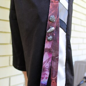 May include: A black and white patterned lanyard with a silver charm attached. The charm is a shield with a design on it. The lanyard is hanging from a person's belt loop.