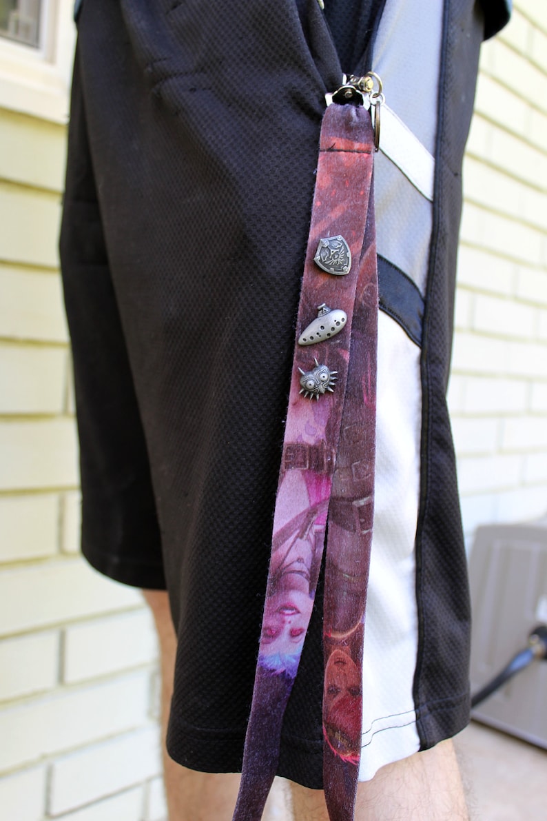 May include: A black and white patterned lanyard with a silver charm attached. The charm is a shield with a design on it. The lanyard is hanging from a person's belt loop.