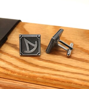 May include: A pair of silver cufflinks with a square design featuring a stylized symbol. The cufflinks are displayed on a wooden box.