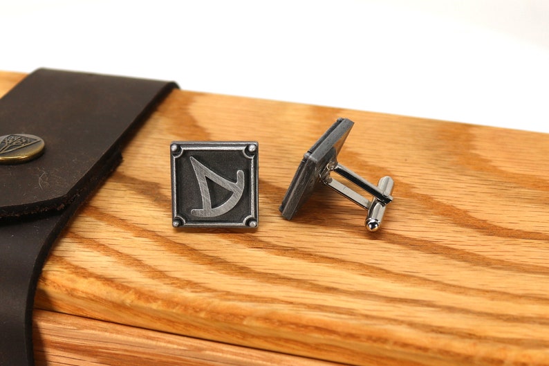 May include: A pair of silver cufflinks with a square design featuring a stylized symbol. The cufflinks are displayed on a wooden box.