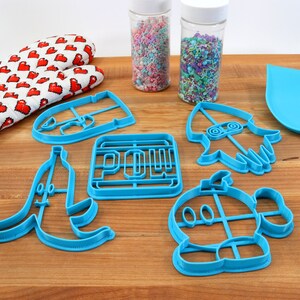 May include: A set of blue plastic cookie cutters shaped like a banana, a video game character, a rocket, a power-up, and a face. The cutters are on a wooden cutting board with a rolling pin, a pair of red oven mitts, and two jars of sprinkles.
