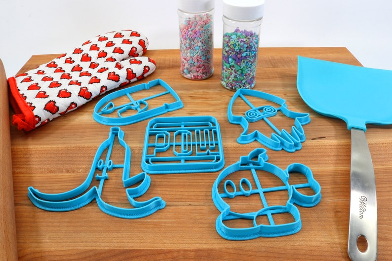May include: A set of blue plastic cookie cutters shaped like a banana, a video game character, a rocket, a power-up, and a face. The cutters are on a wooden cutting board with a rolling pin, a pair of red oven mitts, and two jars of sprinkles.