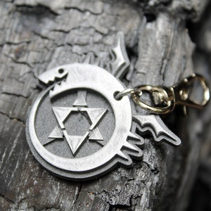 May include: A silver metal keychain featuring a circular design with a star of David inside and a stylized dragon head on the outside. The keychain has a gold metal clasp.