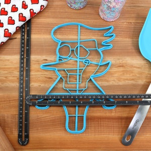 May include: A blue plastic cookie cutter shaped like a cartoon character with glasses and a large nose. The cutter is on a wooden surface with a ruler and a spatula.