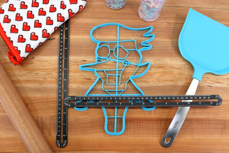 May include: A blue plastic cookie cutter shaped like a cartoon character with glasses and a large nose. The cutter is on a wooden surface with a ruler and a spatula.