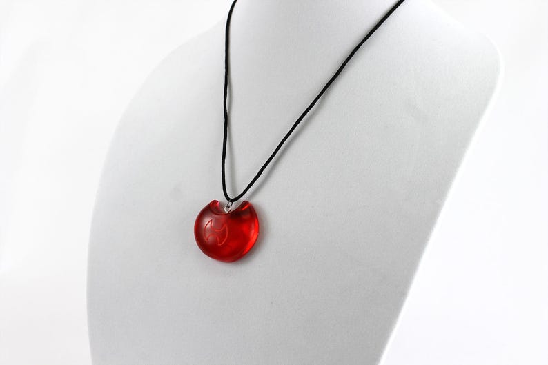 May include: A red heart-shaped pendant necklace with a black cord. The pendant has a silver bail and a small design inside.