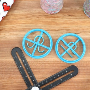 May include: Two blue plastic cookie cutters shaped like a circle with a design inside. The cutters are on a wooden surface.