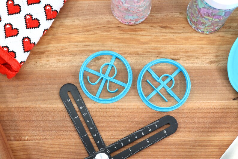May include: Two blue plastic cookie cutters shaped like a circle with a design inside. The cutters are on a wooden surface.