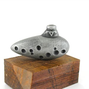 Puede incluir: Una ocarina plateada con un s&iacute;mbolo triangular en la parte superior, sentada sobre una base de madera.