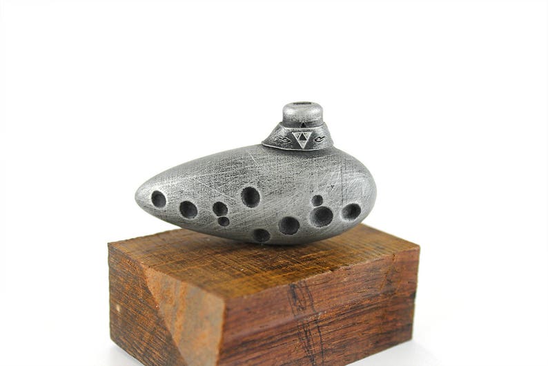 Puede incluir: Una ocarina plateada con un s&iacute;mbolo triangular en la parte superior, sentada sobre una base de madera.