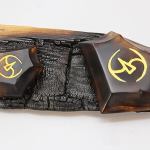 May include: Two brown, geometrically shaped resin pieces with gold accents. The pieces are sitting on a piece of charred wood.