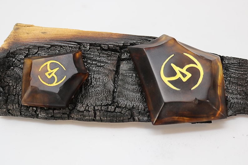 May include: Two brown, geometrically shaped resin pieces with gold accents. The pieces are sitting on a piece of charred wood.