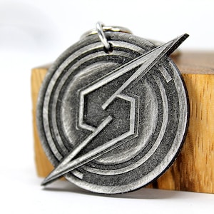 Puede incluir: Un llavero de metal plateado con un logotipo estilizado con un diseño de rayo. El logotipo está rodeado de círculos concéntricos.