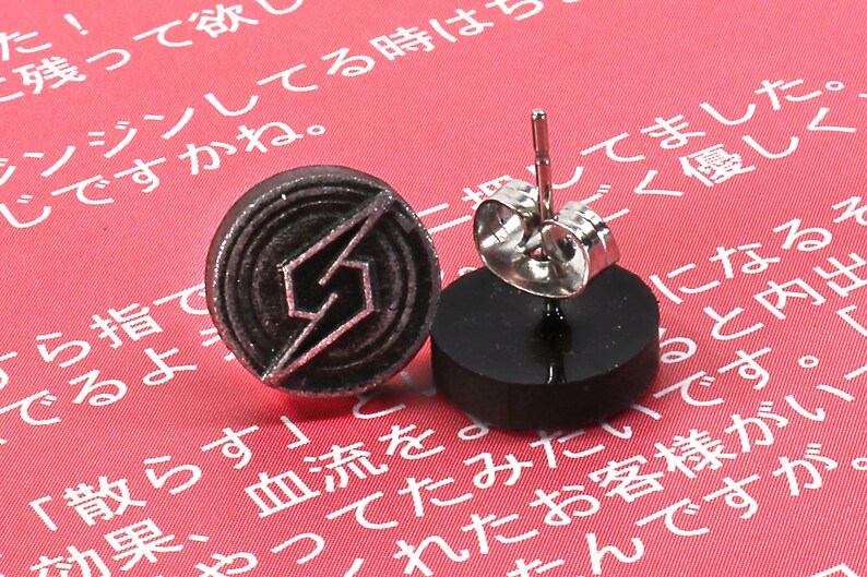 May include: A pair of silver stud earrings with a black circular design featuring a stylised lightning bolt. The earrings are displayed on a red surface.