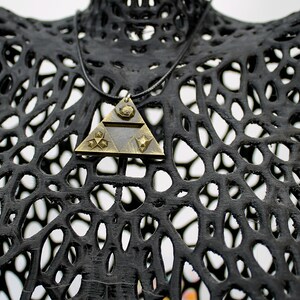 May include: A gold-toned triangular pendant necklace with a black cord. The pendant features a geometric design with a star and a triangle inside. The necklace is displayed on a black, 3D printed mannequin.