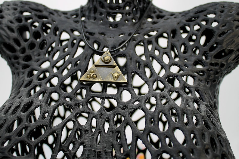 May include: A gold-toned triangular pendant necklace with a black cord. The pendant features a geometric design with a star and a triangle inside. The necklace is displayed on a black, 3D printed mannequin.