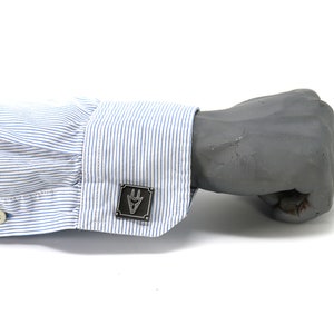 May include: A pair of cufflinks with a silver finish and a geometric design. The cufflinks are on a blue and white striped shirt cuff.