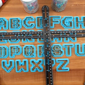 May include: A set of blue plastic alphabet cookie cutters in a pixelated font style. The cutters are arranged on a wooden surface with a black ruler and a blue spatula.