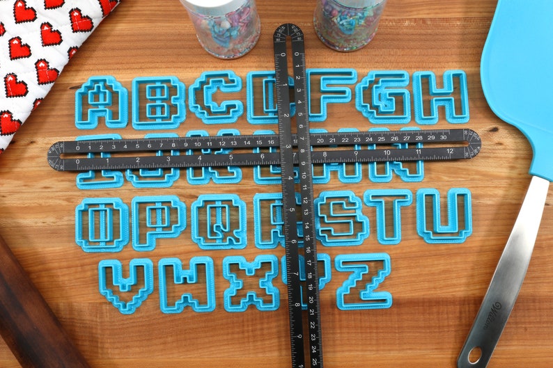 May include: A set of blue plastic alphabet cookie cutters in a pixelated font style. The cutters are arranged on a wooden surface with a black ruler and a blue spatula.