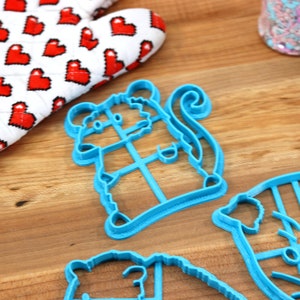 May include: A set of four blue plastic cookie cutters shaped like a mouse, a cat, a bear, and a pig. The cutters are on a wooden surface.