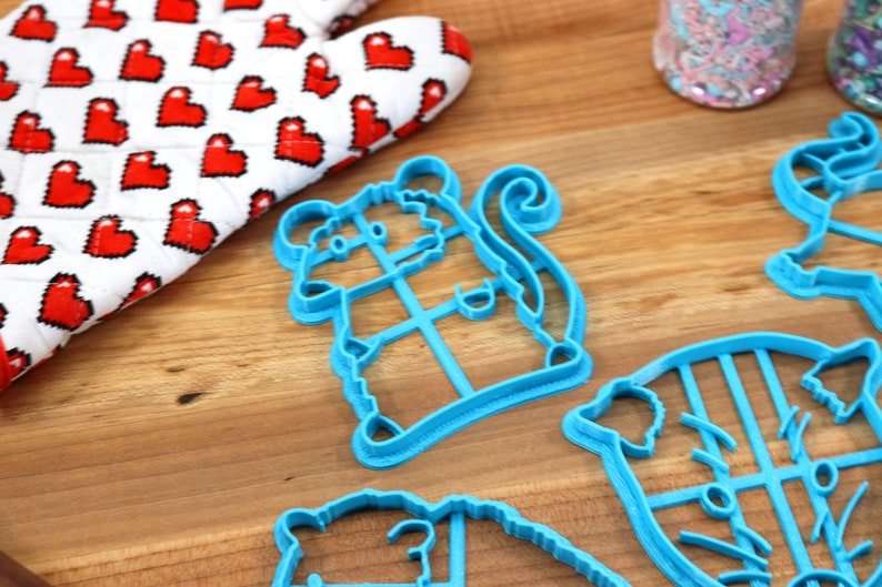 May include: A set of four blue plastic cookie cutters shaped like a mouse, a cat, a bear, and a pig. The cutters are on a wooden surface.