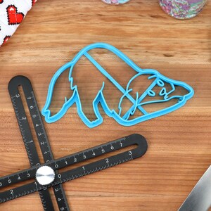 May include: A blue plastic cookie cutter shaped like a sleeping sloth. The cutter is on a wooden surface with a black ruler and a blue spatula.