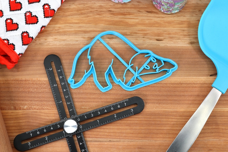 May include: A blue plastic cookie cutter shaped like a sleeping sloth. The cutter is on a wooden surface with a black ruler and a blue spatula.