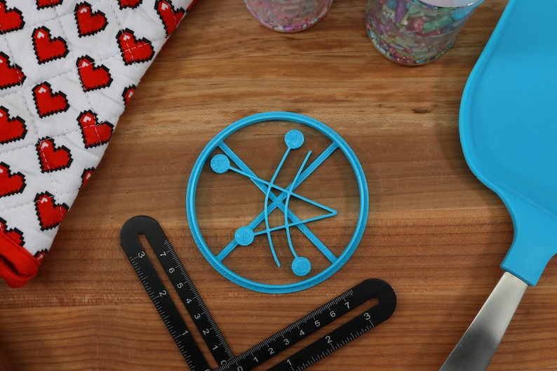 May include: A blue plastic circle cookie cutter with a design of four circles connected by lines. The cookie cutter is on a wooden surface.