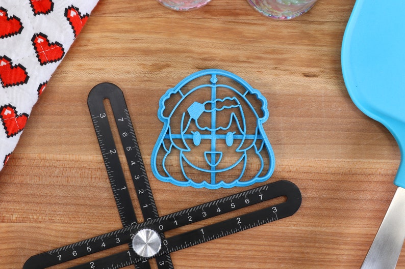 Puede incluir: Cortador de galletas de pl&aacute;stico azul en forma de personaje de dibujos animados con una cara sonriente y cabello largo. El cortador tiene un patr&oacute;n de cuadr&iacute;cula en el interior.